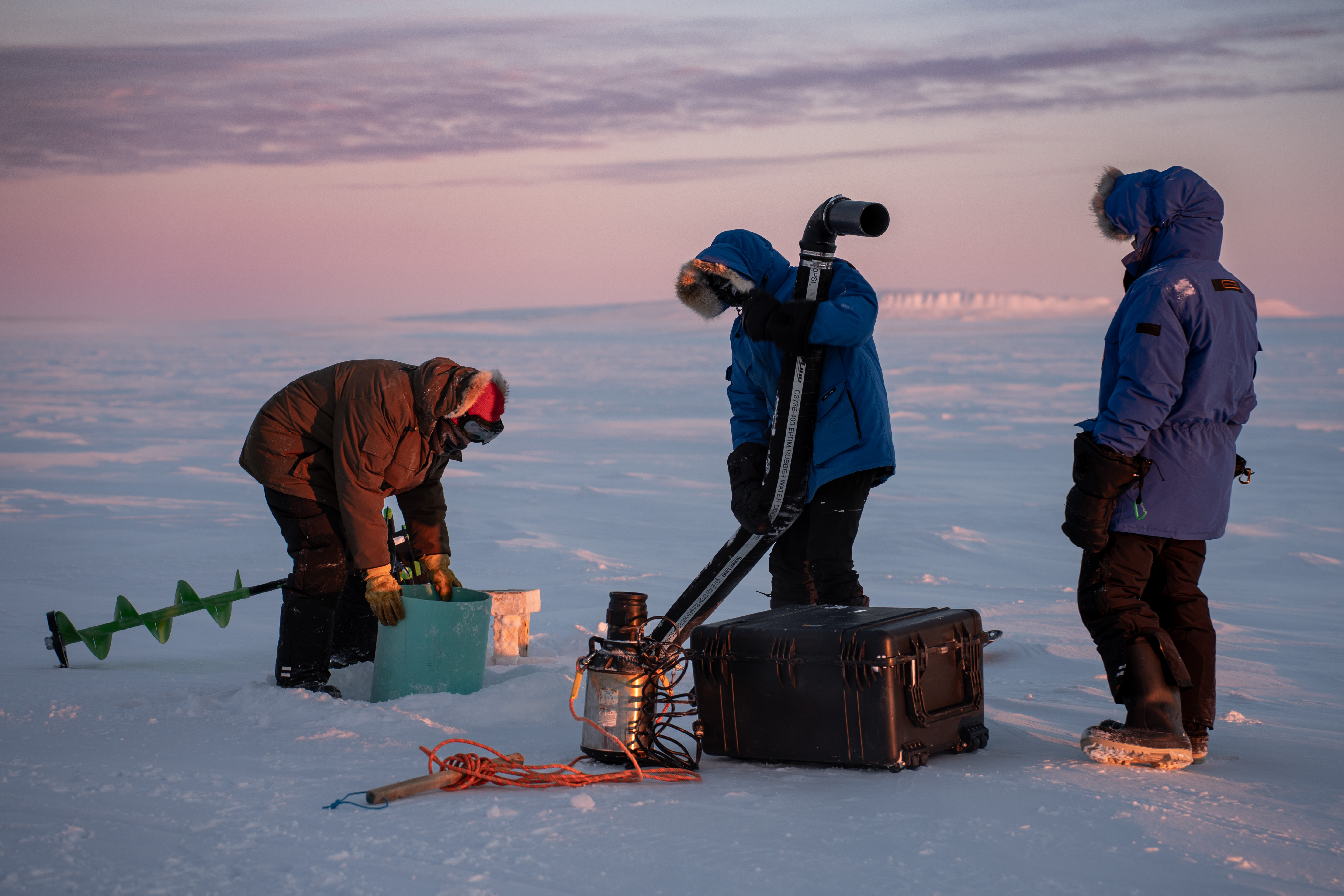A team of three people dressed in warm winter clothes working on a water pump on ice plains in Canada