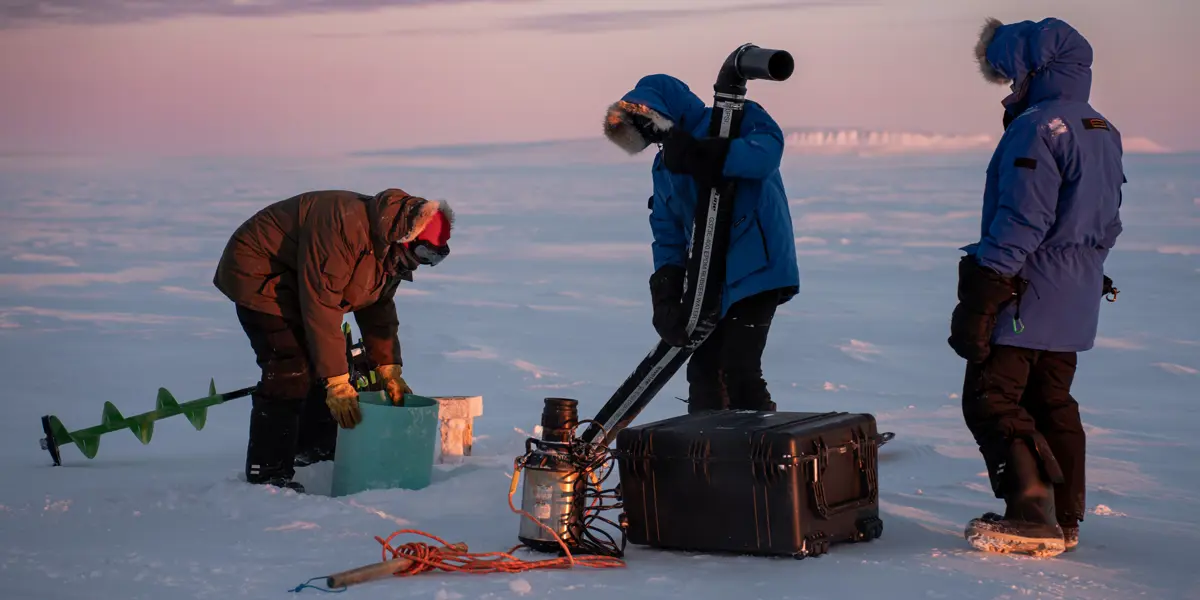 A team of three people dressed in warm winter clothes working on a water pump on ice plains in Canada