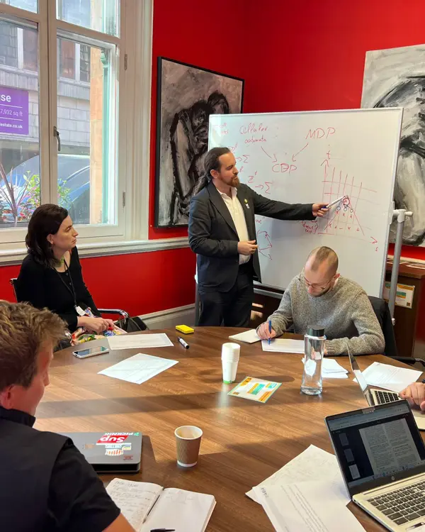 A photo of a group of people sitting around the table and looking at davidad writing on a whiteboard.